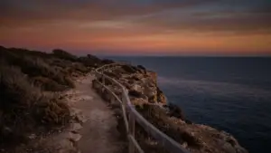 A cliff edge path above a calm sea at dusk with a safety rail