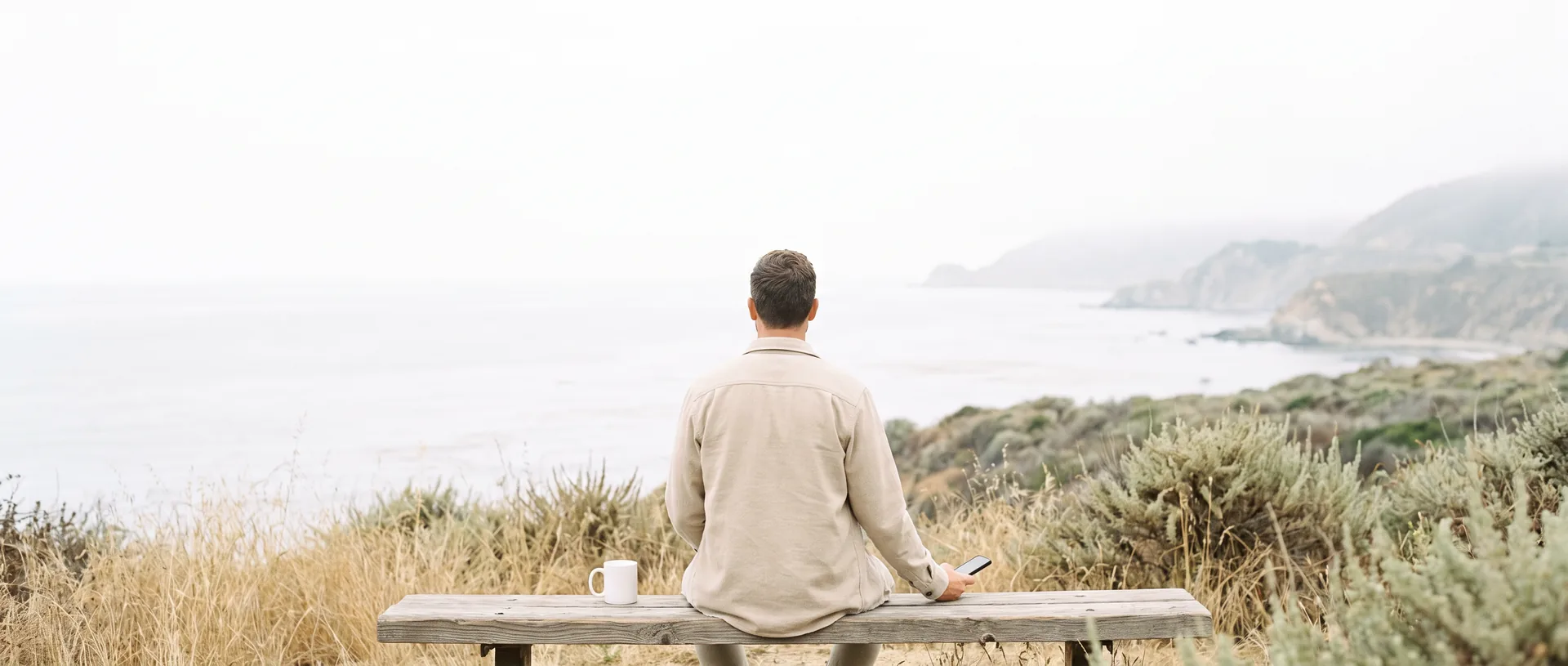 A man seen from behind on a wooden bench above the Pacific on an overcast morning, a phone held loosely at his side and a mug beside him