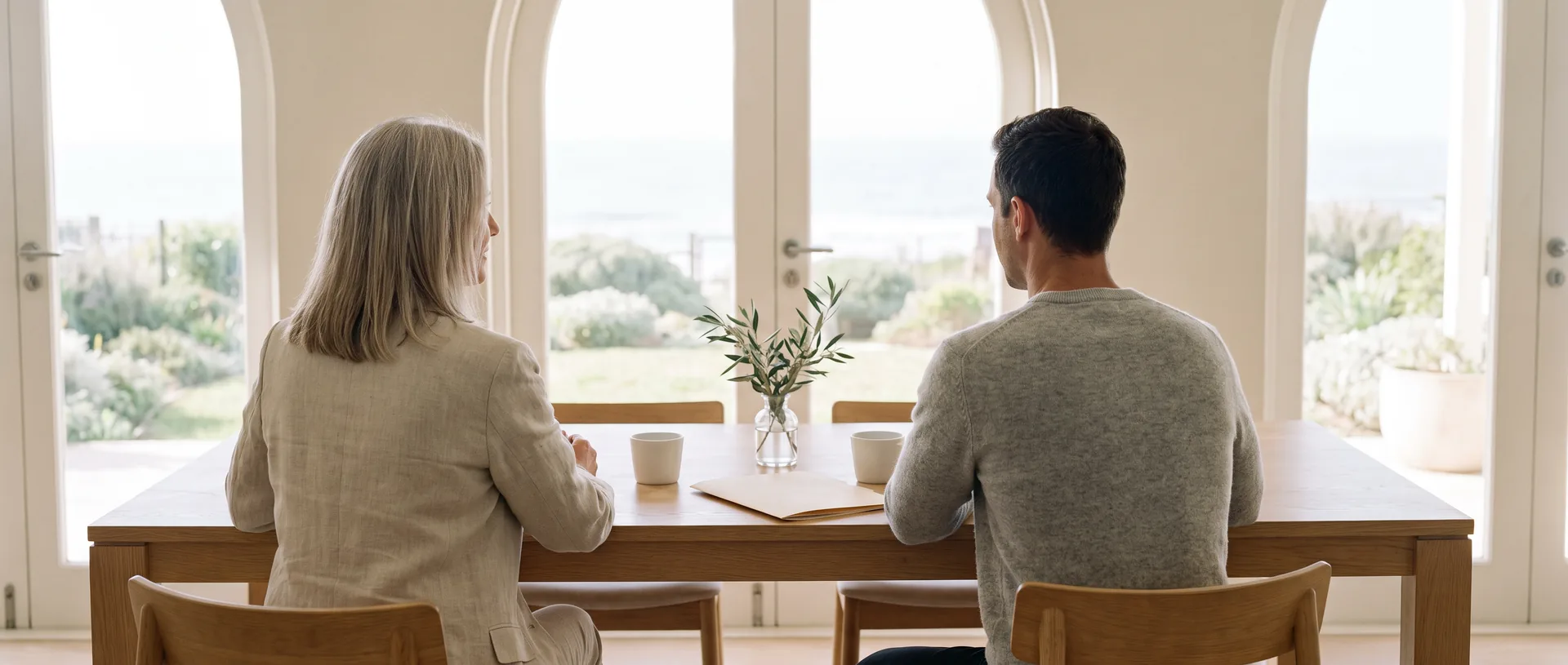 Two colleagues seen from behind at a pale oak table, a closed folder and two coffee cups in front of them, tall arched windows looking out to a coastal garden