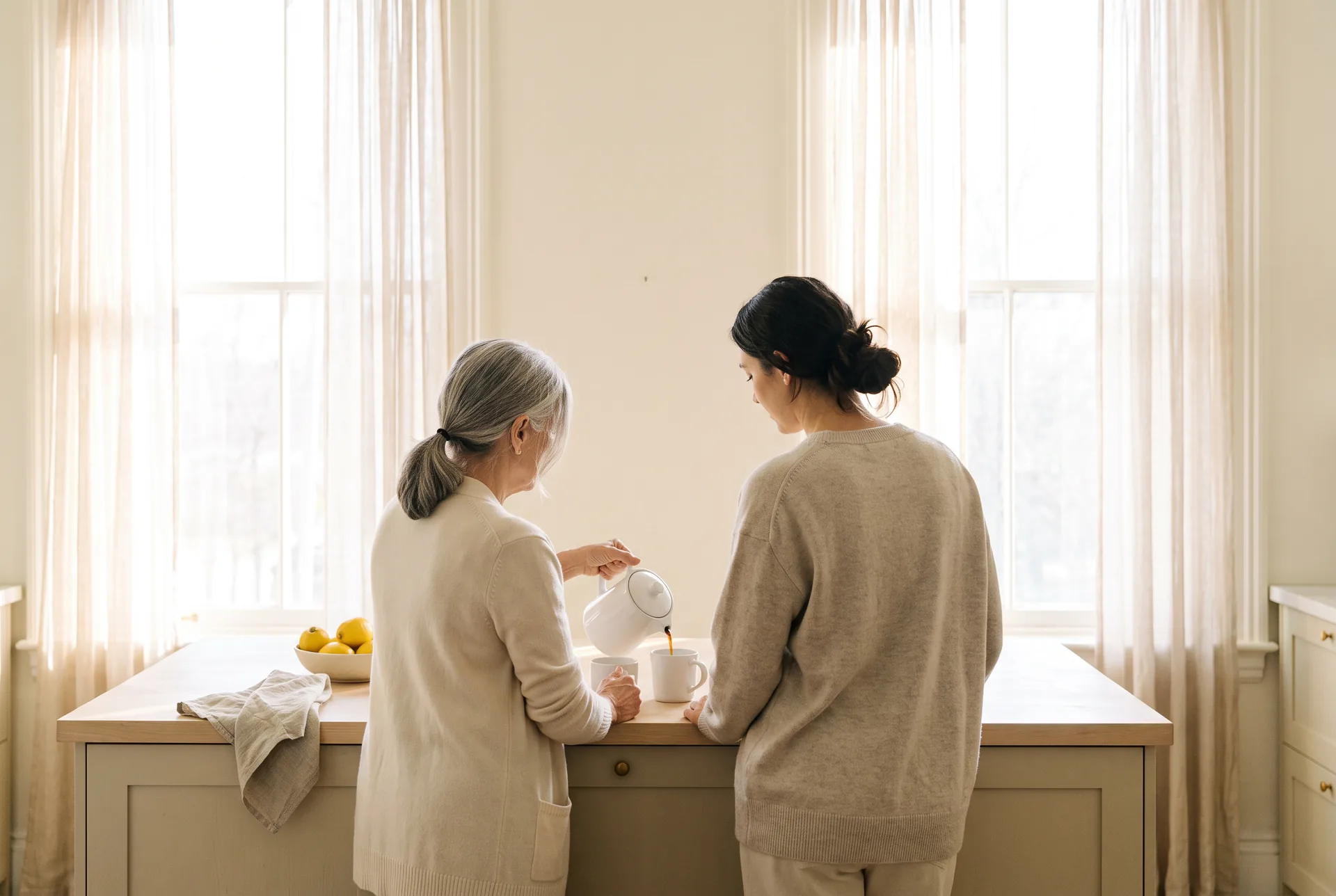 A mother and her adult daughter seen from behind at a kitchen counter, the mother pouring coffee into two mugs