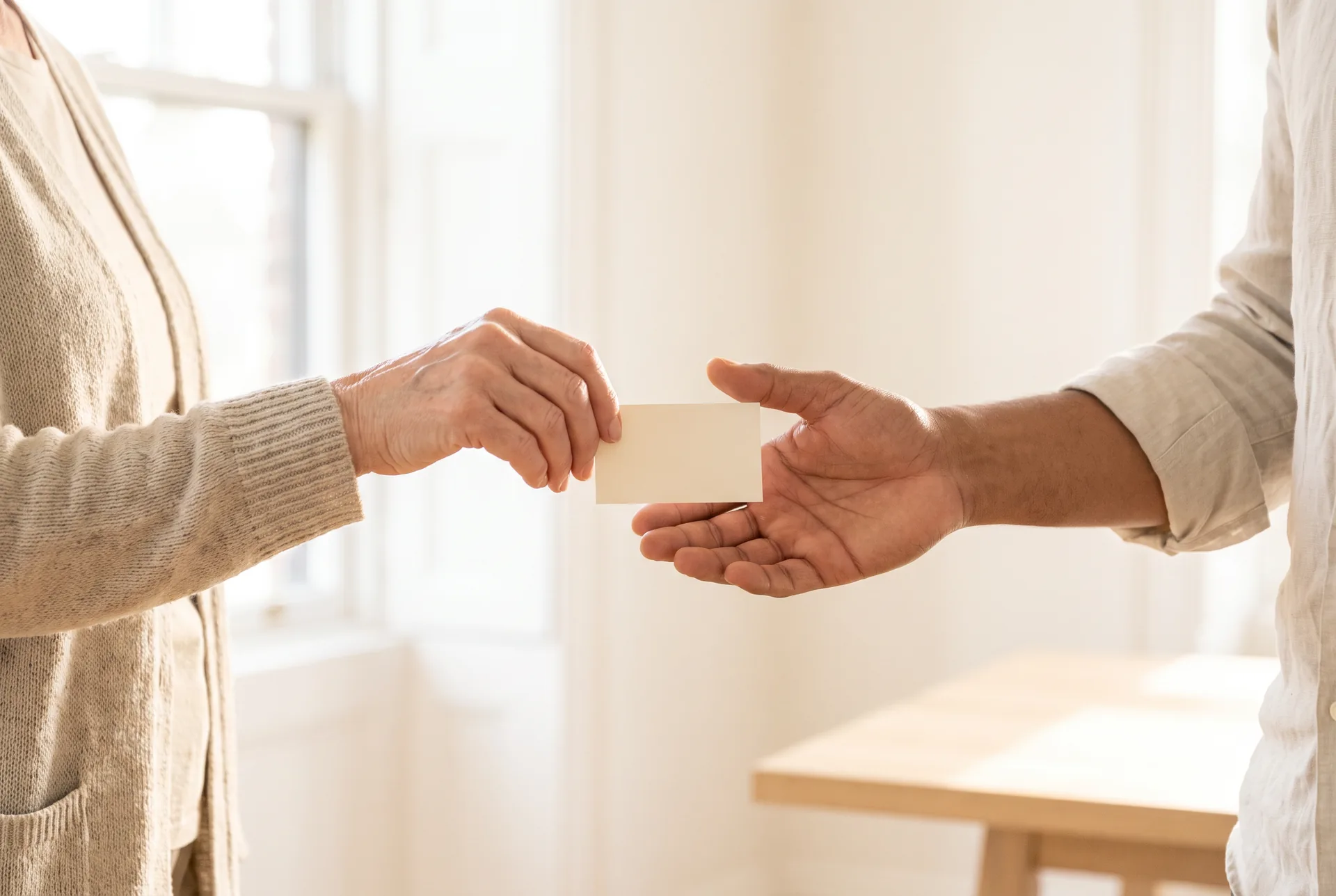One person's hand passing a small blank card to another person's open hand in warm window light